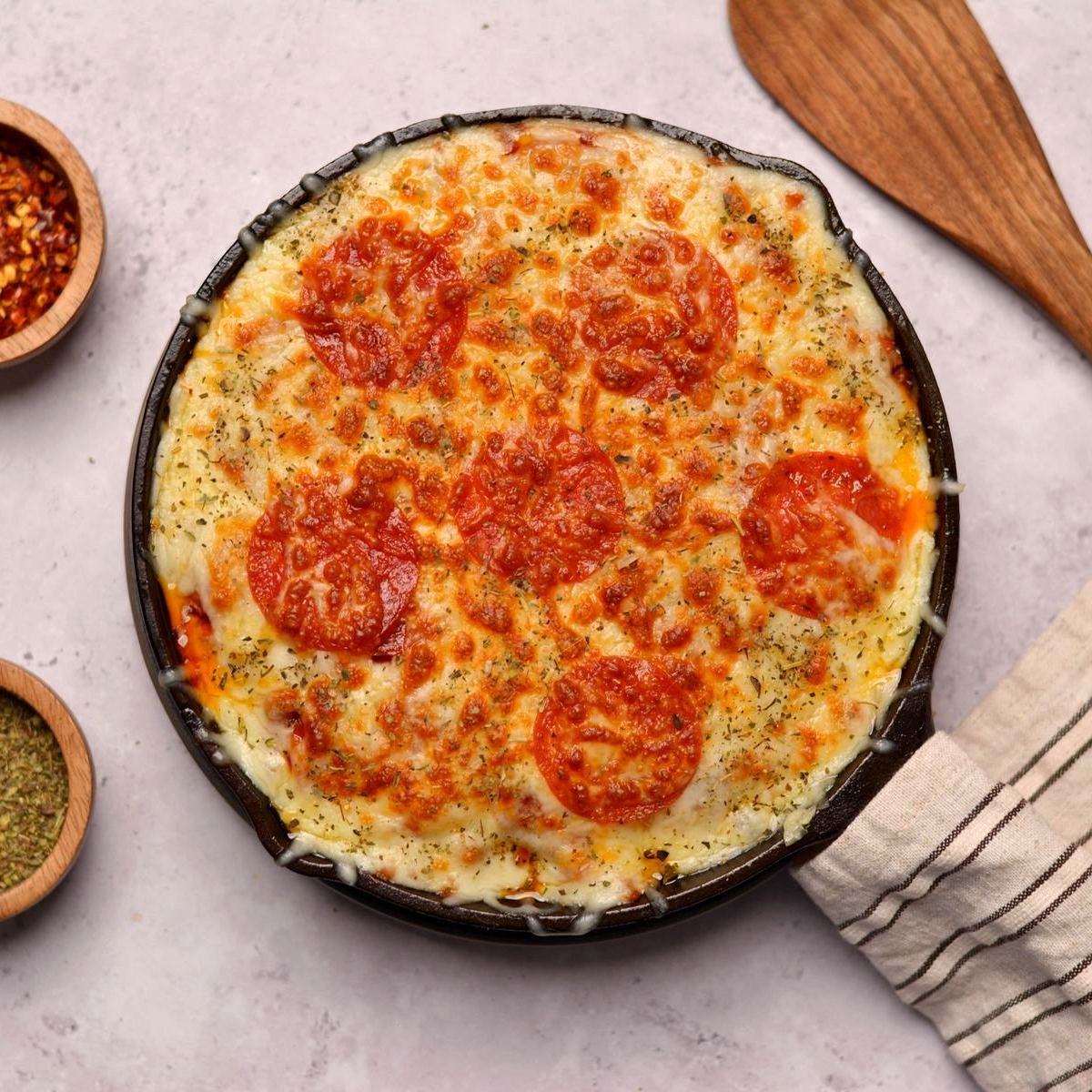 Pepperonis and golden brown cheese in a black skillet with spices in wooden bowls and a wooden spoon on the side.