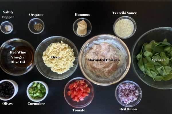 Ingredients for Gyro bowl laid out and labeled with salt, pepper, oregano, hummus, tzatziki sauce, spinach, marinated chicken, orzo, red wine vinegar, olive oil, olives, cucumber, tomato, and red onion