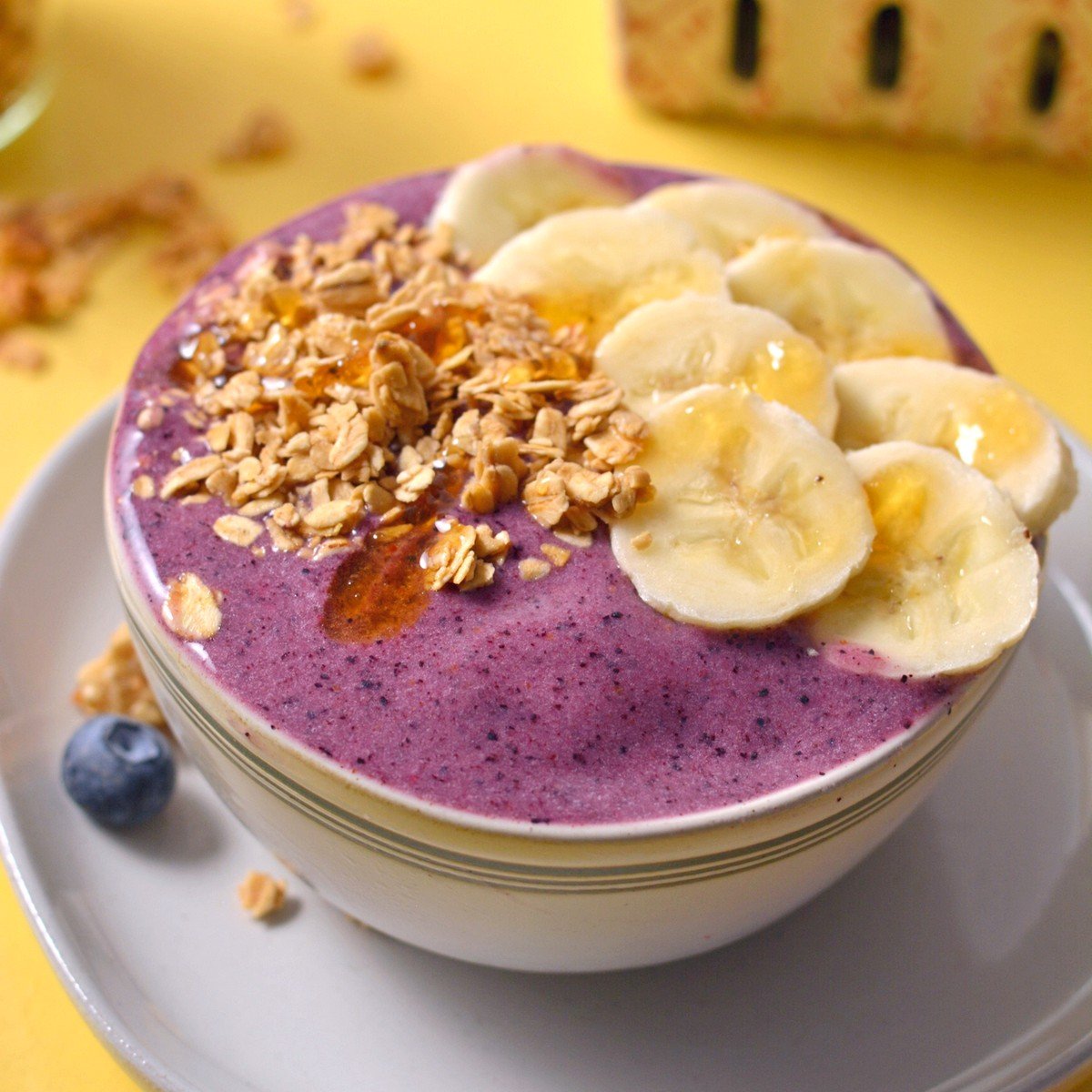 Purple smoothie bowl with bananas and granola on top and a yellow background.
