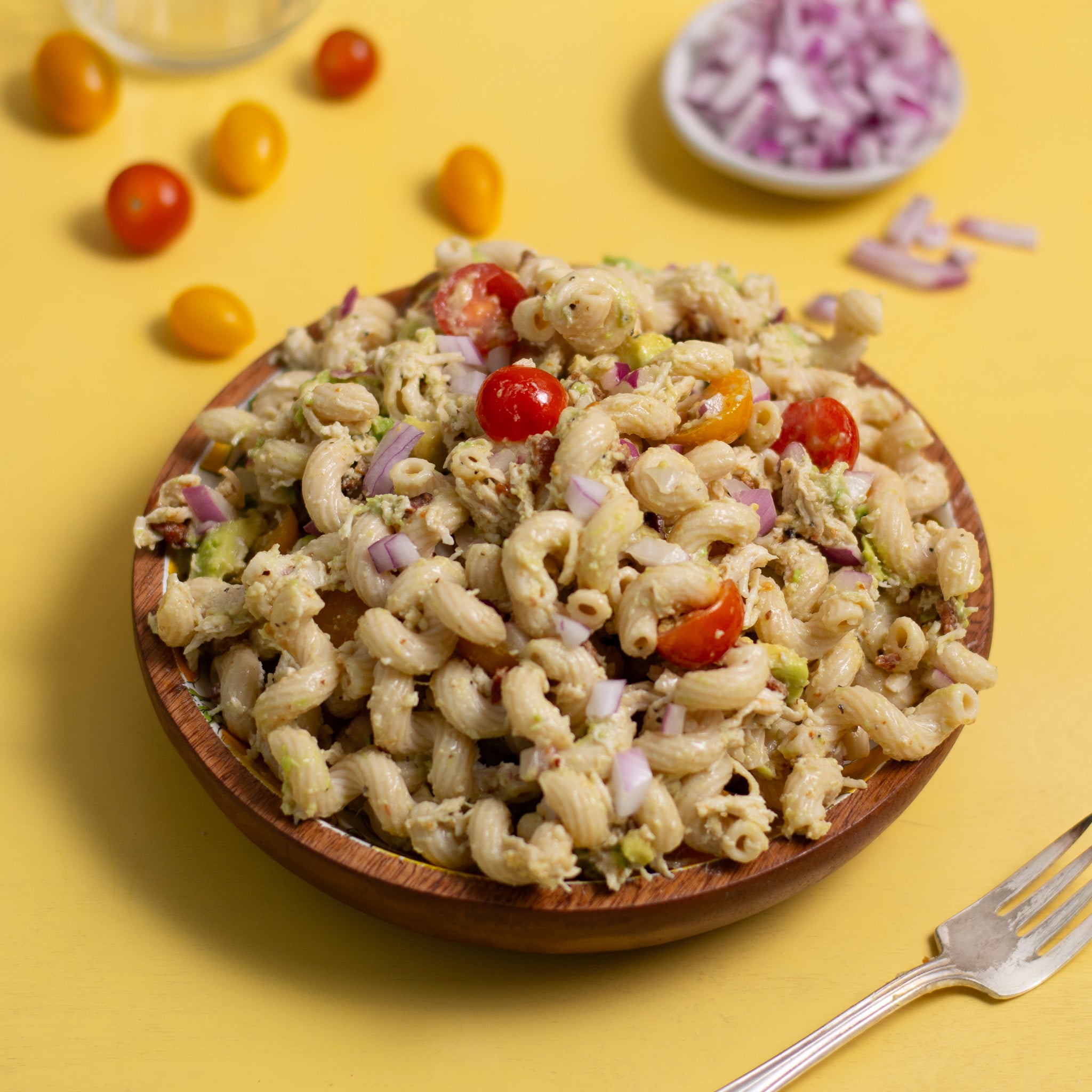 A wooden bowl filled with creamy protein BLT pasta salad rests on a yellow surface. The salad, featuring elbow macaroni, cherry tomatoes, red onion bits, and green herbs, delights the senses. In the background, chopped onions, grape tomatoes, and a fork are visible.