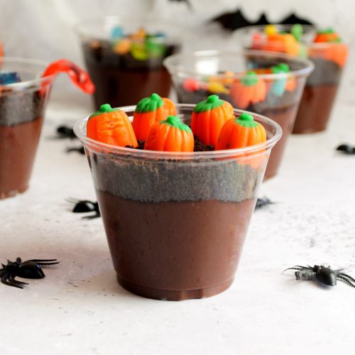 Chocolate pudding with crushed oreos and pumpkin candy on top inside a plastic cup.