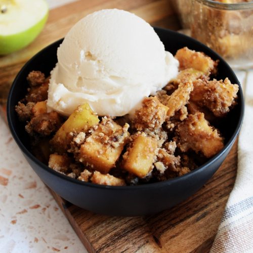 Apple cobbler in a black bowl with vanilla ice cream on top.