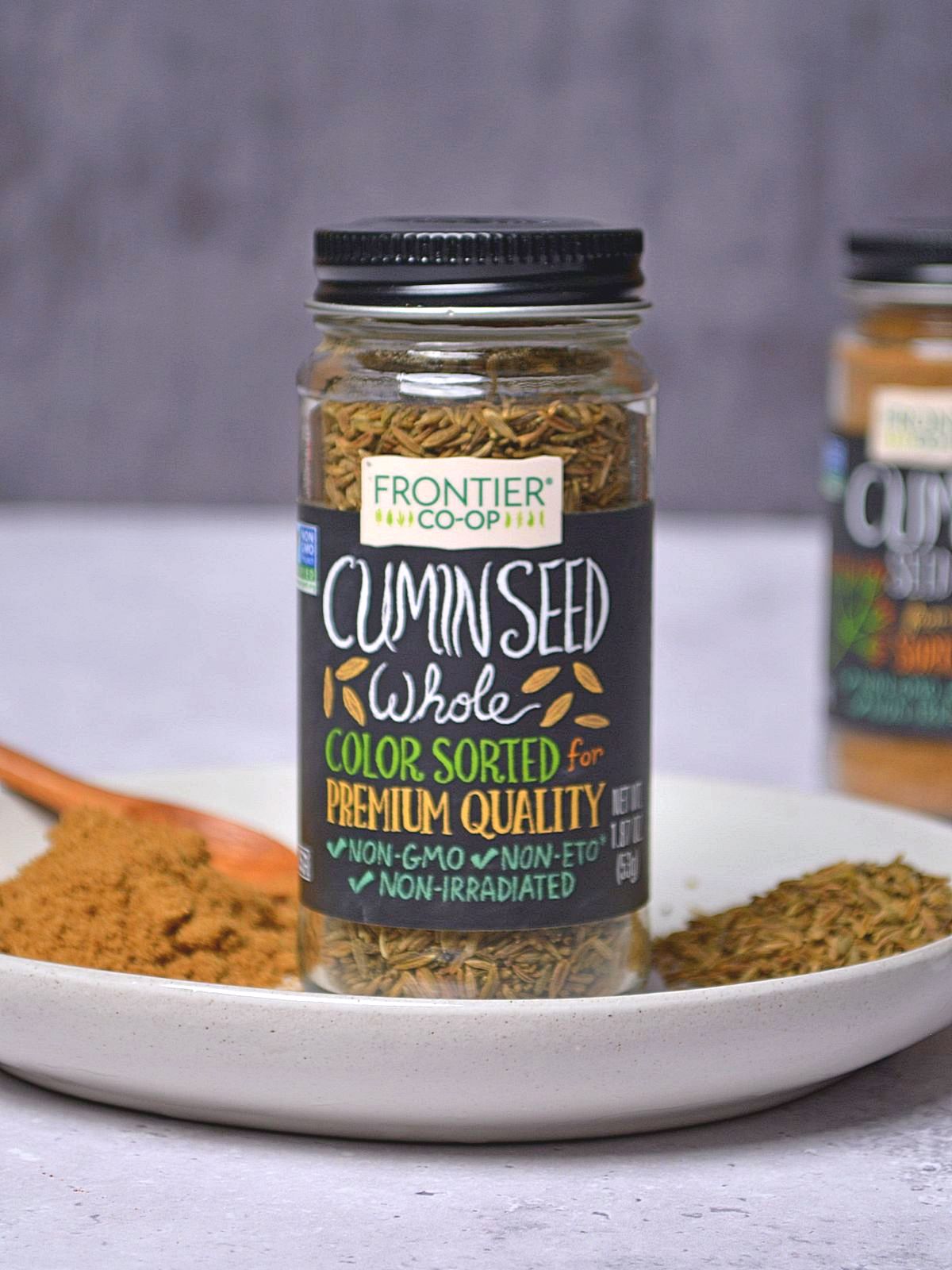 Cumin in a spice jar set on a white plate with ground cumin on the plate