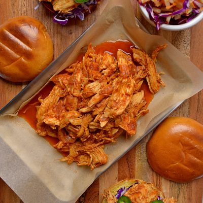 Pulled chicken on a dish over parchment paper with a sandwich and cole slaw next to it
