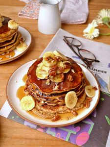 Stack of pancakes with bananas and pecans on top on a white plate set on top of a newspaper with glasses, flowers, and more pancakes in the background.