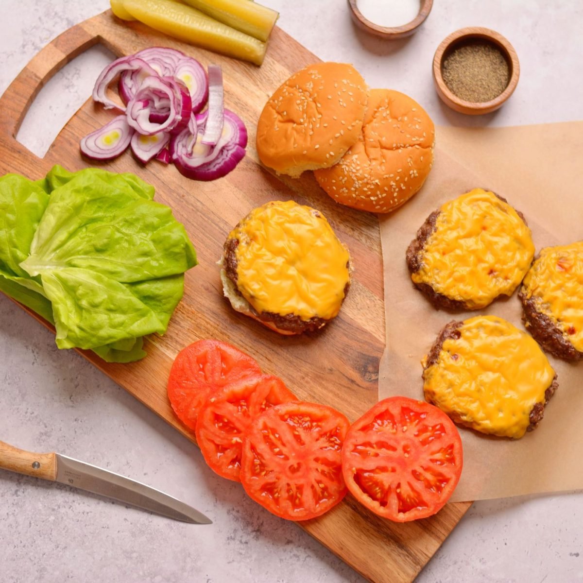 Burgers with cheese, tomaotes, lettuce, red onion, pickles on a wood cutting board.