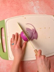 Hands using a knife to chop a red onion on a white cutting board.