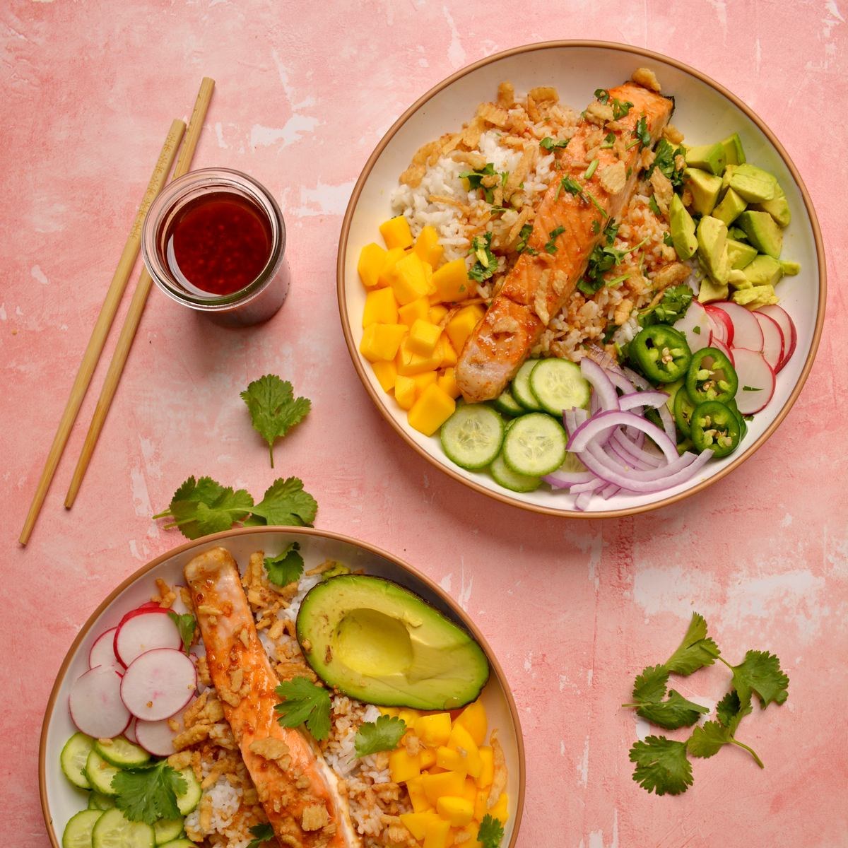 Two shallow bowls filled with rice, salmon, and various fruits and vegetables.