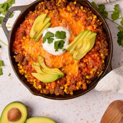 Flat beef enchiladas in a black skillet with avocado and sour cream on top.