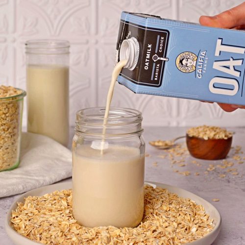 Oat milk being poured into a glass mason jar surrounded by oats.