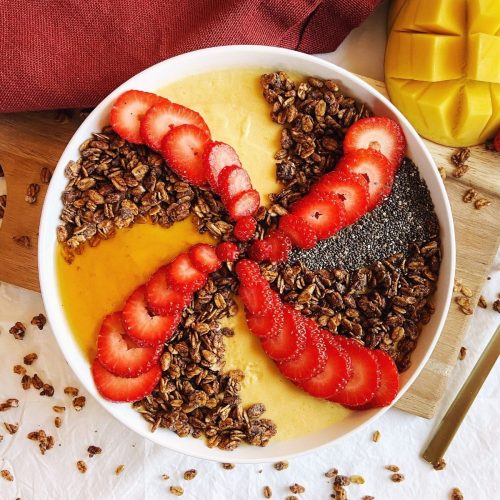 A mango smoothie bowl with strawberry slices and granola in a swirl shape.