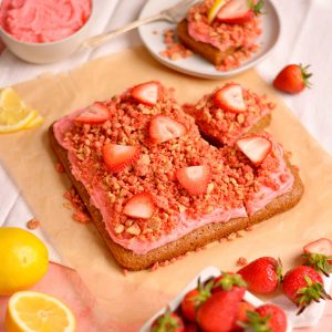A square cake topped with red frosting, red crunch, and strawberry slices.