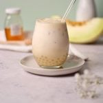 A glass of creamy honeydew milk tea with a glass straw sits on a saucer. Tapioca pearls are visible at the bottom. In the background, there’s a jar of honey and a slice of melon. The setting is on a light-colored surface.