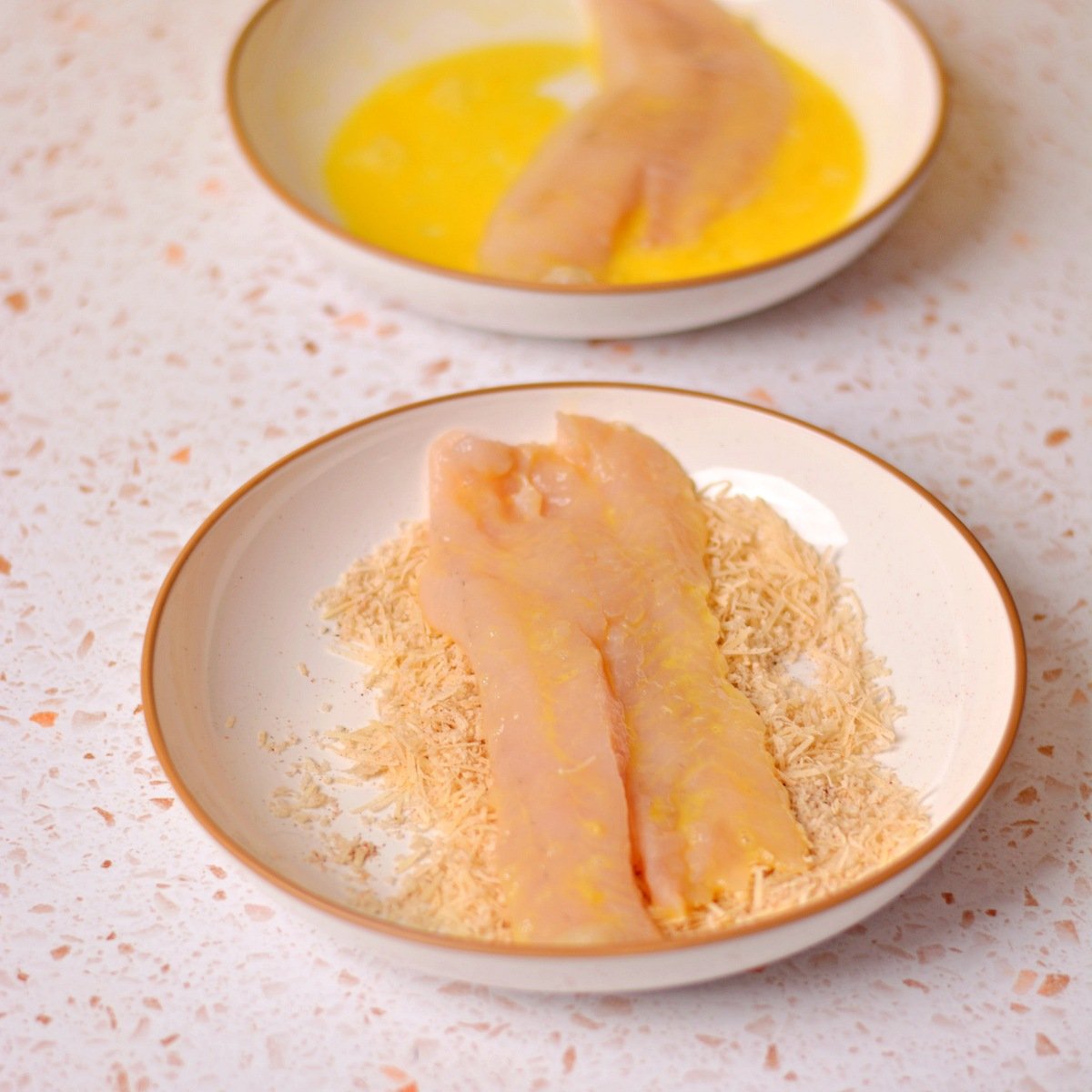 A fish fillet in a bowl with egg and one in a bowl with breadcrumbs.