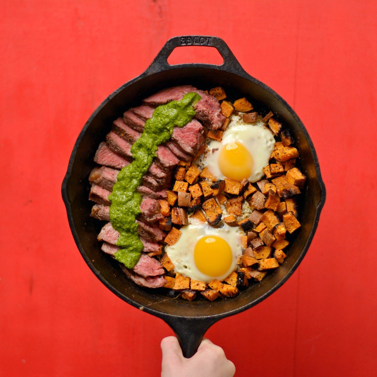 A hand holding the skillet of potatoes, eggs, and steak.