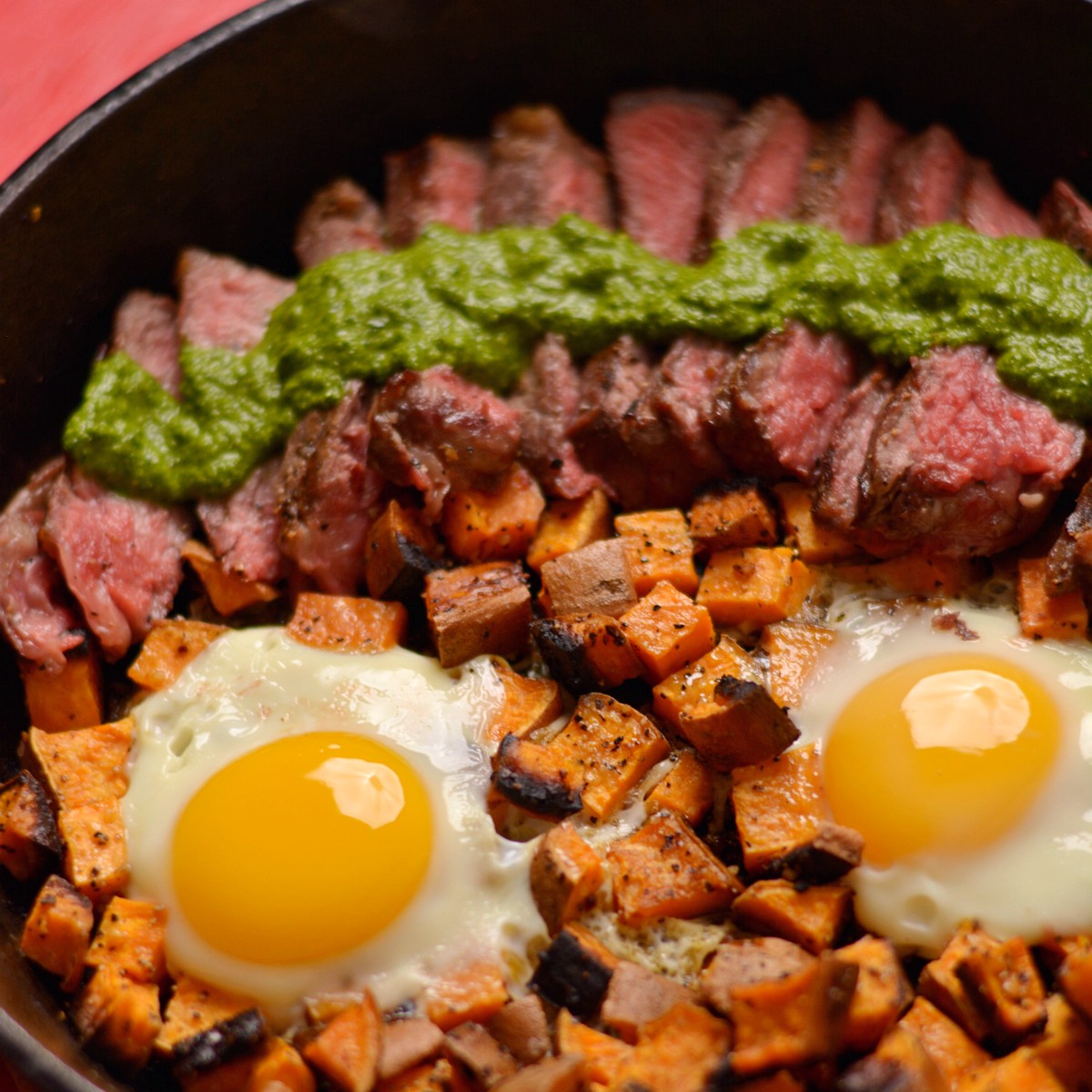 A skillet with steak, sweet potatoes, and eggs with a green sauce.