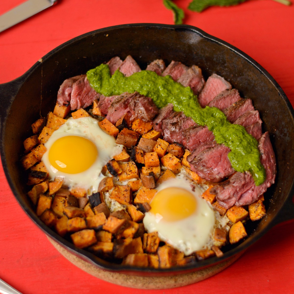 A skillet with steak, chimichurri sauce, eggs, and potatoes.