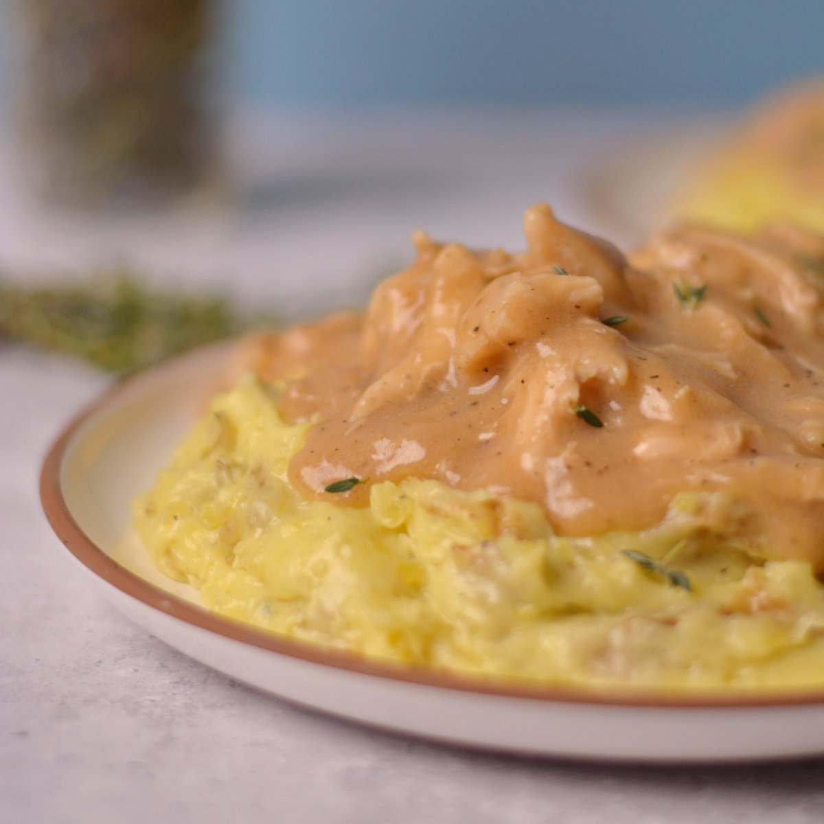 Mashed potatoes on a plate with chicken and gravy on top and thyme in the background.