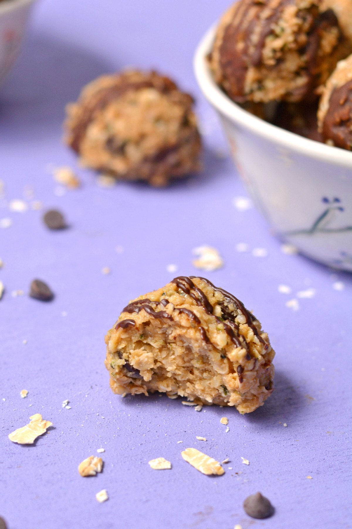 A protein ball with chocolate drizzle and a bite take from in in front of a purple background.