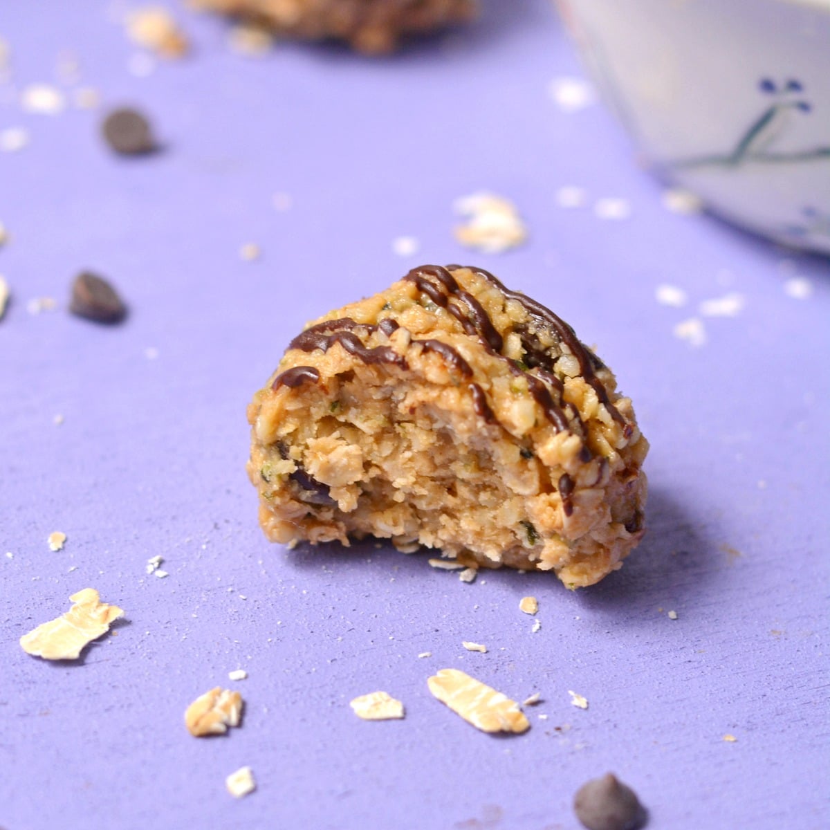 A protein ball with chocolate drizzle and a bite take from in in front of a purple background.