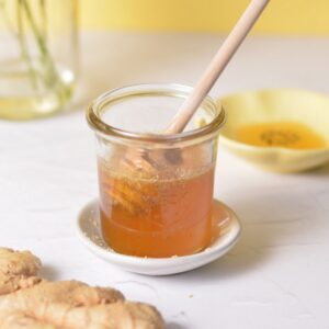 A jar of honey syrup with a honey stick in front of a yellow background.