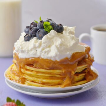 A stack of protein pancakes with greek yogurt on top and nut butter syrup on a purple background.
