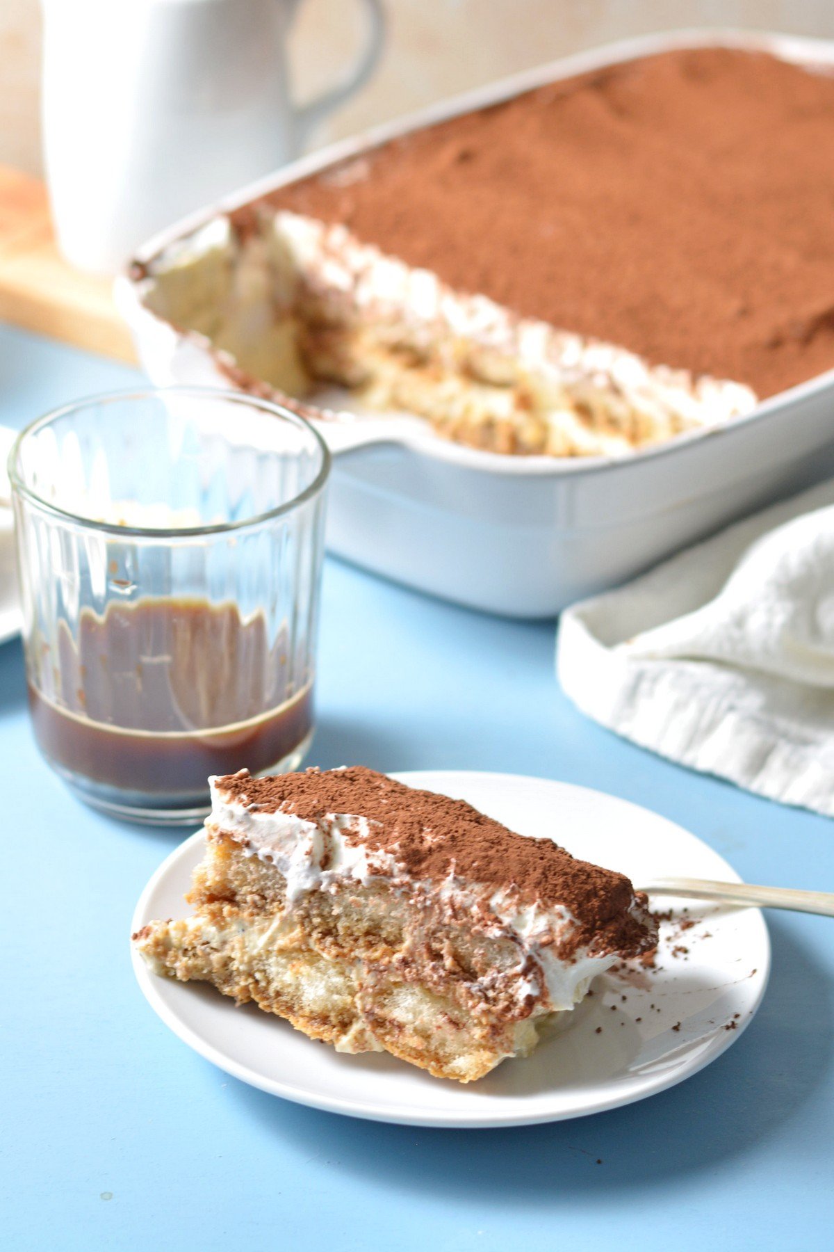Protein tiramisu on a plate in front of a cup of coffee and a tray of the dessert.