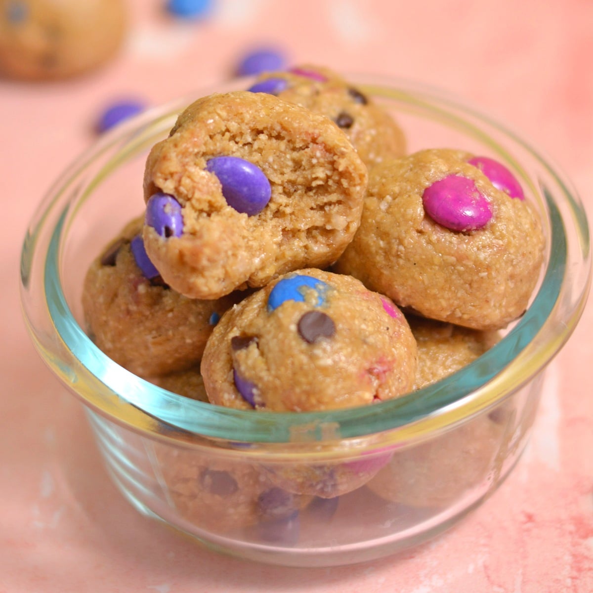 A glass bowl filled with monster cookie protein bites, studded with colorful candies and chocolate chips. One bite is taken out of the top piece, revealing the soft texture inside. The background features soft pink tones sprinkled with a few candies.