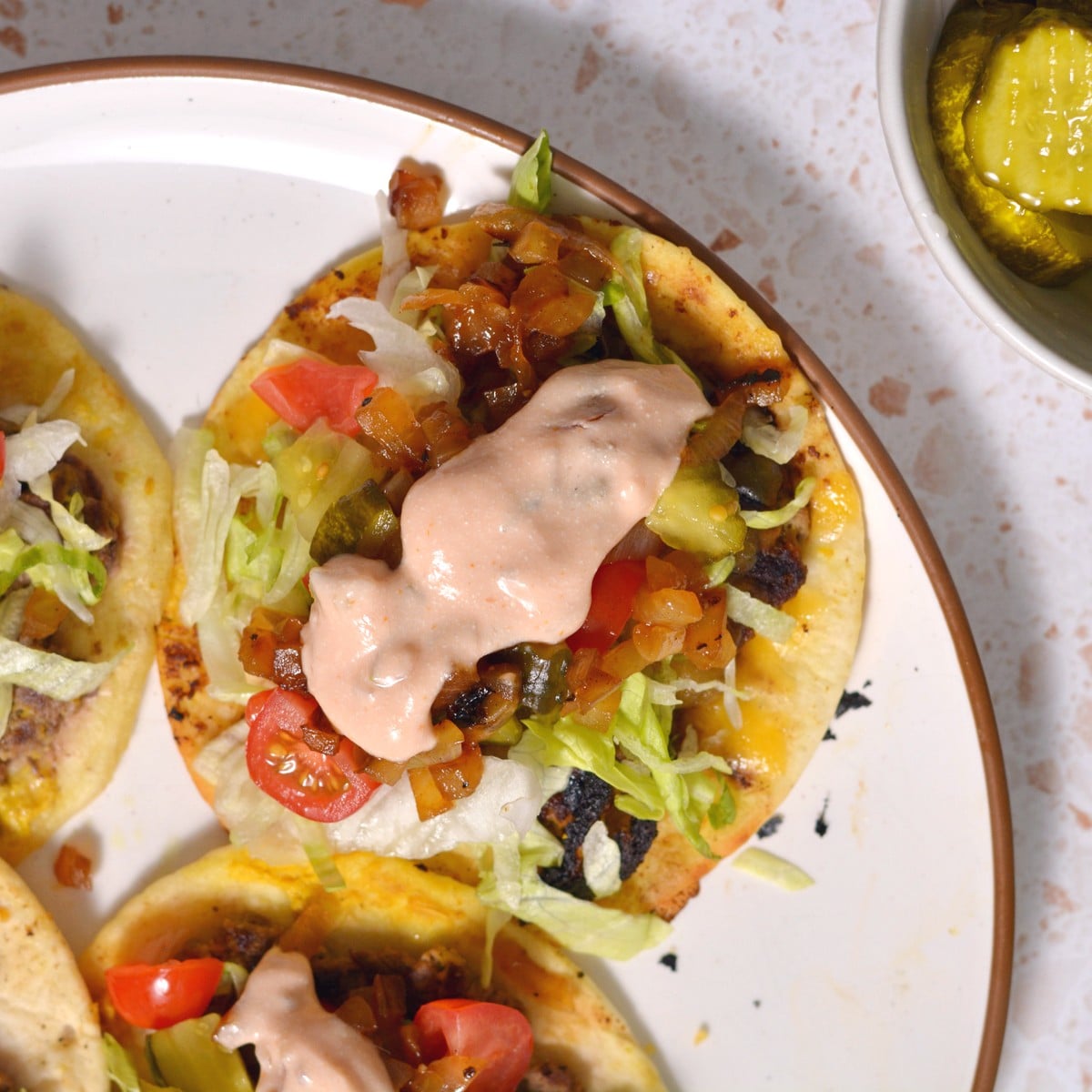 Close-up of a plate with high-protein tacos topped with shredded lettuce, diced tomatoes, grilled vegetables, and a dollop of special creamy sauce. A small bowl of pickles is in the background on a speckled surface.