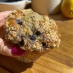 A high protein blueberry muffin inside a paper muffin cup with a hand holding it.
