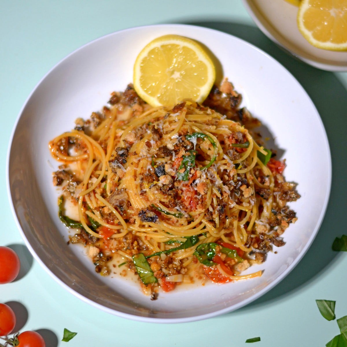 A plate of bone broth pasta topped with a savory crumb mixture, cherry tomatoes, fresh herbs, and a lemon slice. The dish is set on a light green surface with extra lemon and tomatoes nearby.