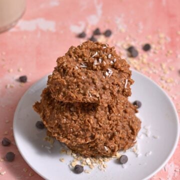Three no-bake protein cookies stacked on a white plate, sprinkled with shredded coconut and surrounded by oats and chocolate chips, on a pink surface.