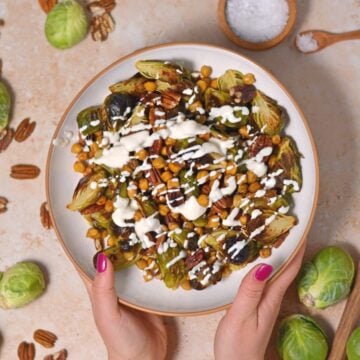 A white bowl filled with brussels sprouts, chickpeas, and pecans with a protein greek yogurt sauce drizzled on top.
