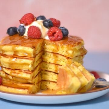 A stack of fluffy protein pancakes topped with whipped cream, raspberries, and blueberries, drizzled with syrup. A fork with a bite-sized piece rests on the plate beside these delicious pancakes.