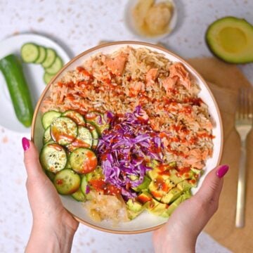 A white bowl with salmon and rice, cucumbers, cabbage, avocado, and ginger that's drizzled with sriracha sauce.