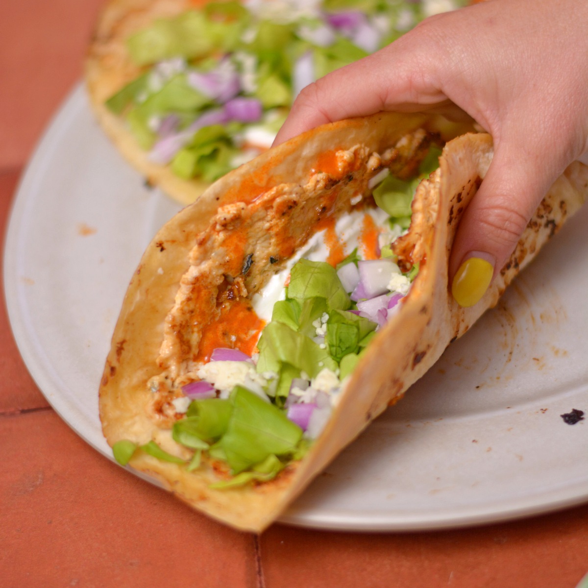 A hand with yellow nail polish holds a Chicken Taco filled with lettuce, diced red onion, creamy sauce, and orange salsa on a white plate. Another taco is partially visible in the background.