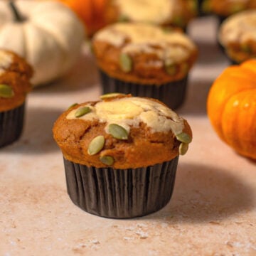 A protein pumpkin muffin topped with pepitas and cream cheese sits on a light surface, surrounded by small orange and white pumpkins in the background.