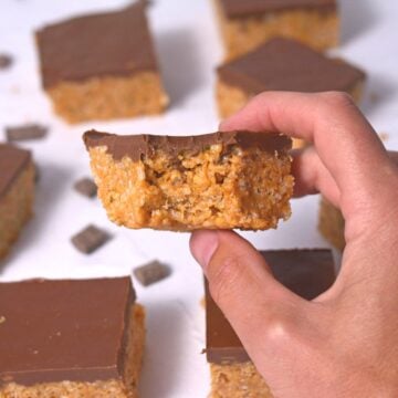 A hand holding a Chocolate Peanut Butter Rice Krispie Bar with a bite taken out, surrounded by more bars and chocolate pieces on a white surface.