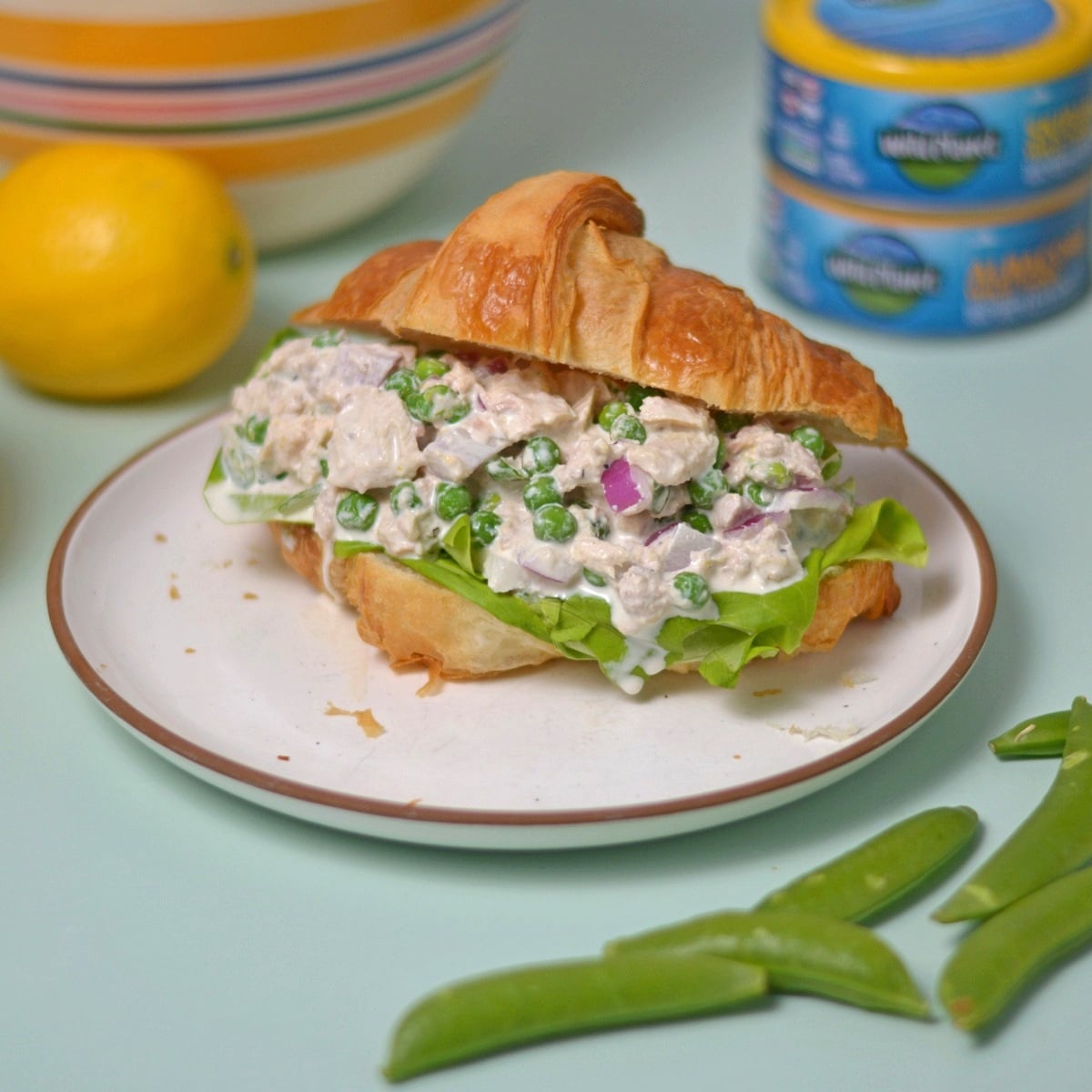 A croissant sandwich filled with tuna salad, green peas, red onions, and lettuce sits on a white plate. Nearby are snap peas, a lemon, a bowl, and stacked cans of tuna for this high protein meal.