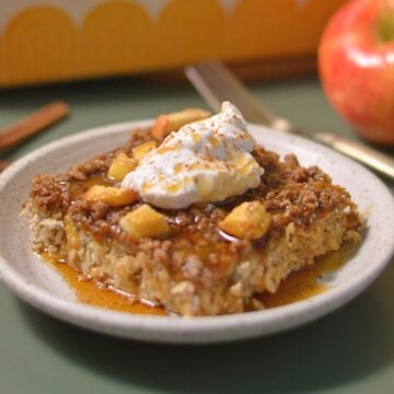 A square piece of baked oatmeal topped with diced apples, syrup, and a dollop of whipped cream sits on a small plate, with an apple and utensils visible in the background.