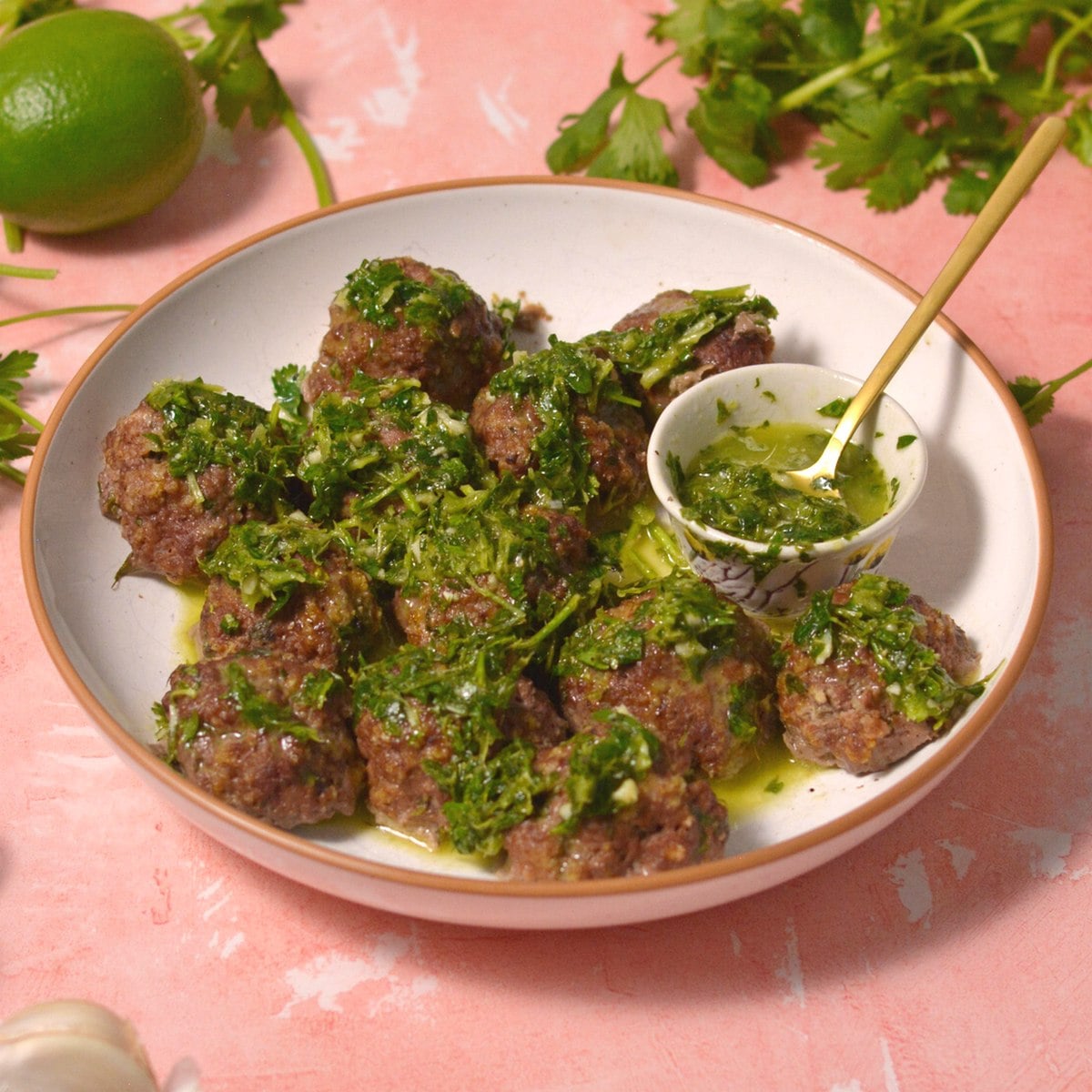 A plate of meatballs topped with green herb sauce sits on a pink surface, with a small dish of extra sauce and a spoon. Fresh herbs and a lime are in the background.