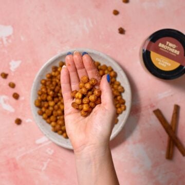 A hand holding roasted chickpeas over a bowl filled with more chickpeas on a pink surface. A jar labeled Two Brothers and cinnamon sticks are also visible nearby.