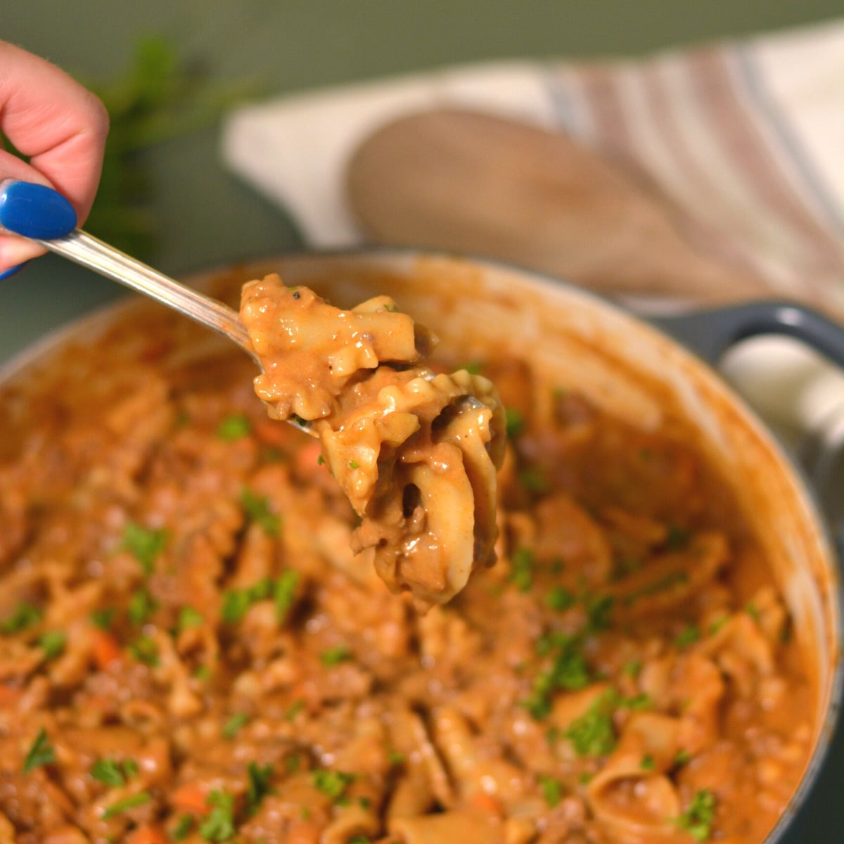 A hand with blue nail polish holds a fork with creamy, saucy pasta above a pot filled with the same pasta dish, garnished with chopped herbs. A striped towel and wooden spoon are in the background.