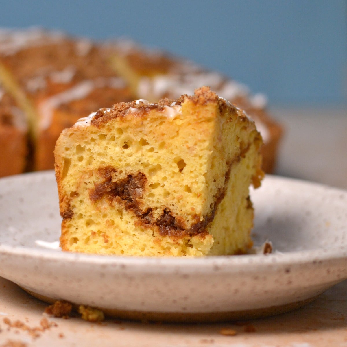 A slice of coffee cake with a cinnamon swirl sits on a speckled ceramic plate. The cake appears moist and fluffy, with a crumb topping and a drizzle of icing. The background shows more of the cake and a light blue wall.