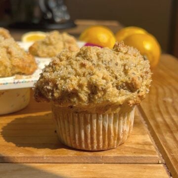 A close-up of a golden-brown Lemon Poppy Seed Protein Muffin with a sugared top sits on a wooden surface, with more muffins and whole lemons blurred in the background. Sunlight highlights the muffins texture.