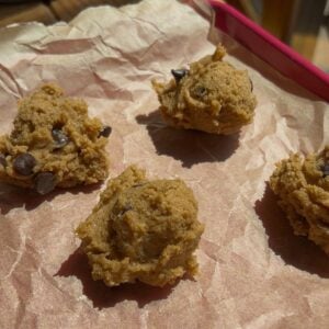 Four scoops of easy protein cookie dough sit on crumpled parchment paper in a pink baking tray, ready to be baked. Sunlight casts warm shadows across the scene.