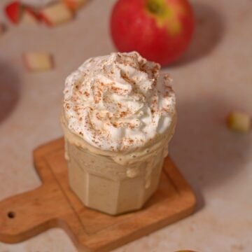 A glass of creamy apple milkshake topped with whipped cream and sprinkled with cinnamon sits on a small wooden board. Apple pieces and a whole apple are in the background.