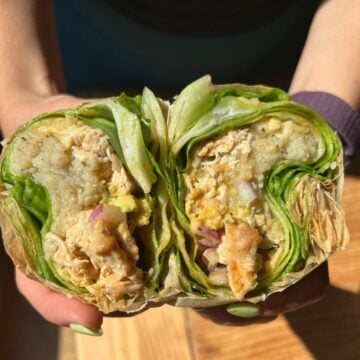 Two hands hold a halved buffalo lettuce wrap filled with rice, chicken, eggs, and onions. The fresh ingredients and texture layers are clearly visible inside the open wrap.