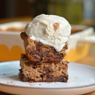 Two thick, chewy blondie bars stacked on a white plate, topped with a scoop of melting vanilla ice cream. A yellow baking dish is visible in the background.