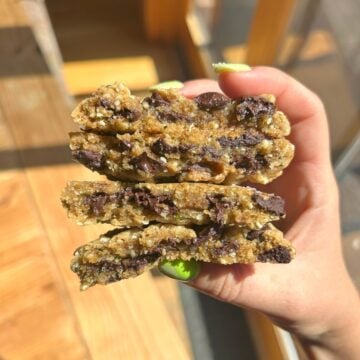 A hand with green painted nails holds a stack of ooey-gooey chocolate chip cookies broken in half, revealing their melty chocolate chips and soft, chewy center. Warm sunlight shines on the cookies and wooden floor below.